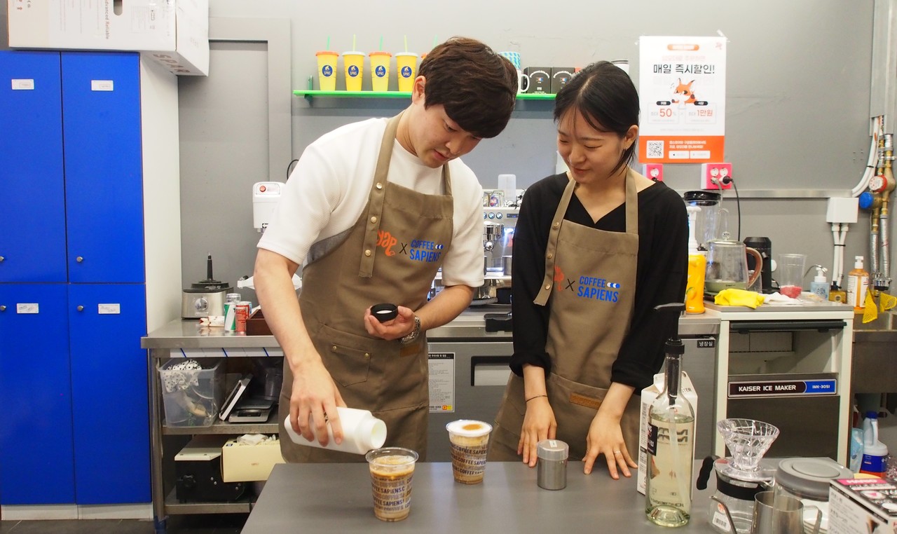 [보도사진] 얍모바일 사업팀 문주영 대리(왼쪽)가 커피사피엔스에서 발굴한 '청년 사피엔스' 김예진 매니저(오른쪽)에게 시그니처 커피 메뉴 솔트크림라떼를 배우고 있다_220530