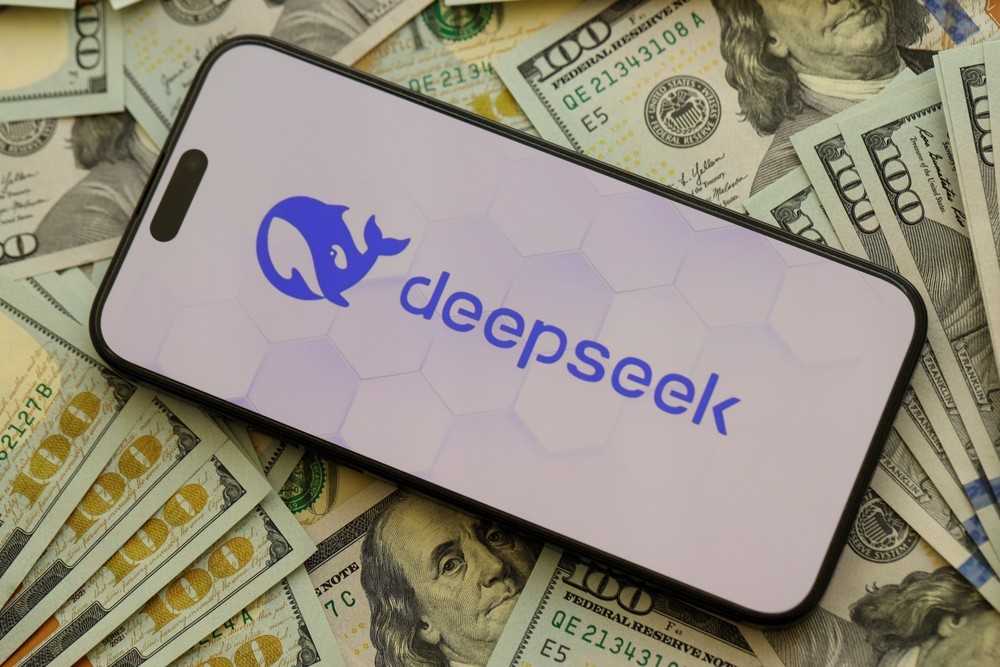 Arka planında dağınık halde ABD doları yüz dolarlık banknotlar bulunan, üzerinde DeepSeek logosu görünen bir akıllı telefon.